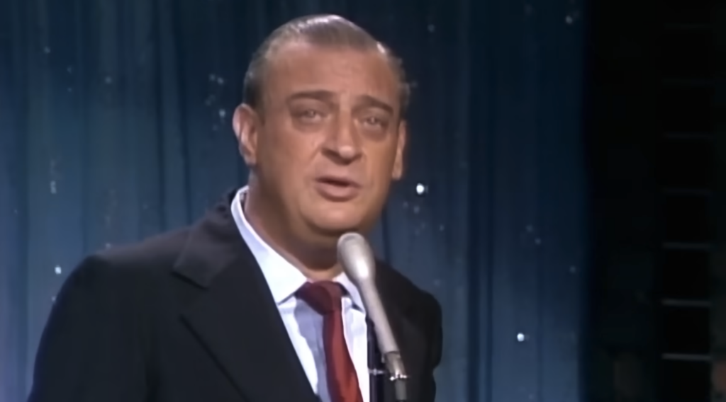 A man with slicked-back gray hair, wearing a dark suit, white shirt, and red tie, stands in front of a microphone on a stage with a dark blue curtain backdrop. He appears to be speaking or performing.