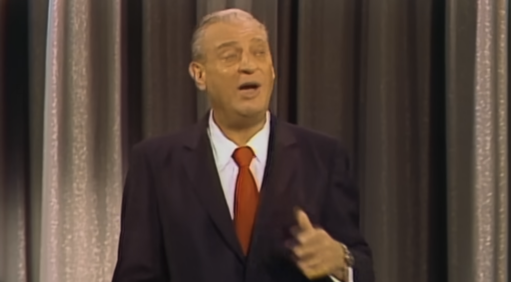 A person wearing a dark suit, white shirt, and red tie stands in front of a curtain, gesturing with one hand and looking ahead. The background curtain alternates between dark and light vertical stripes.