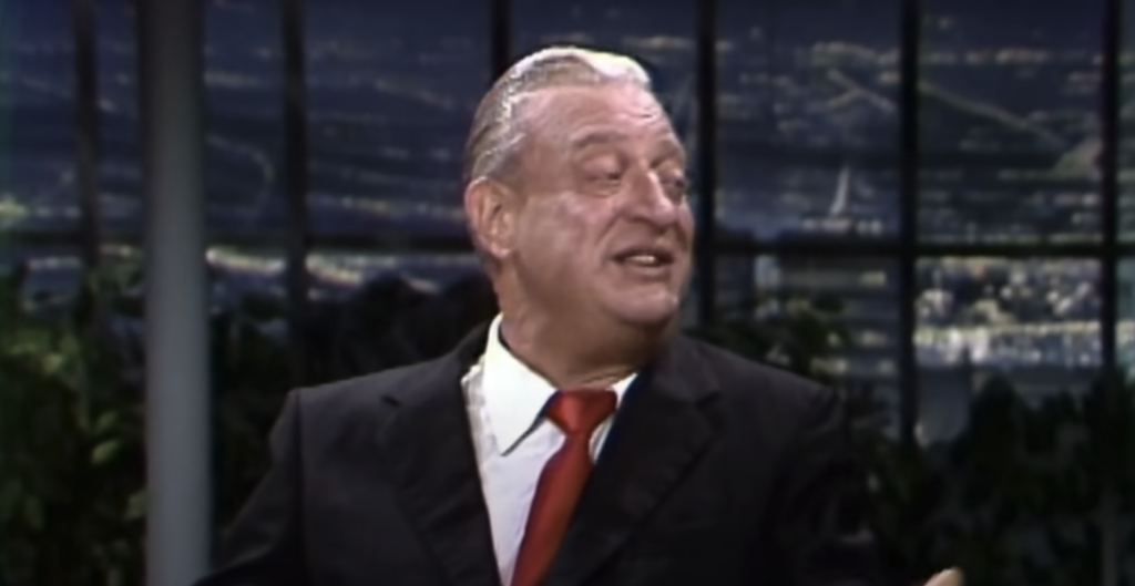 A man with slicked-back gray hair, wearing a black suit, white shirt, and red tie, is speaking passionately on a night-time talk show set. The background features large windows showcasing a cityscape illuminated by lights.