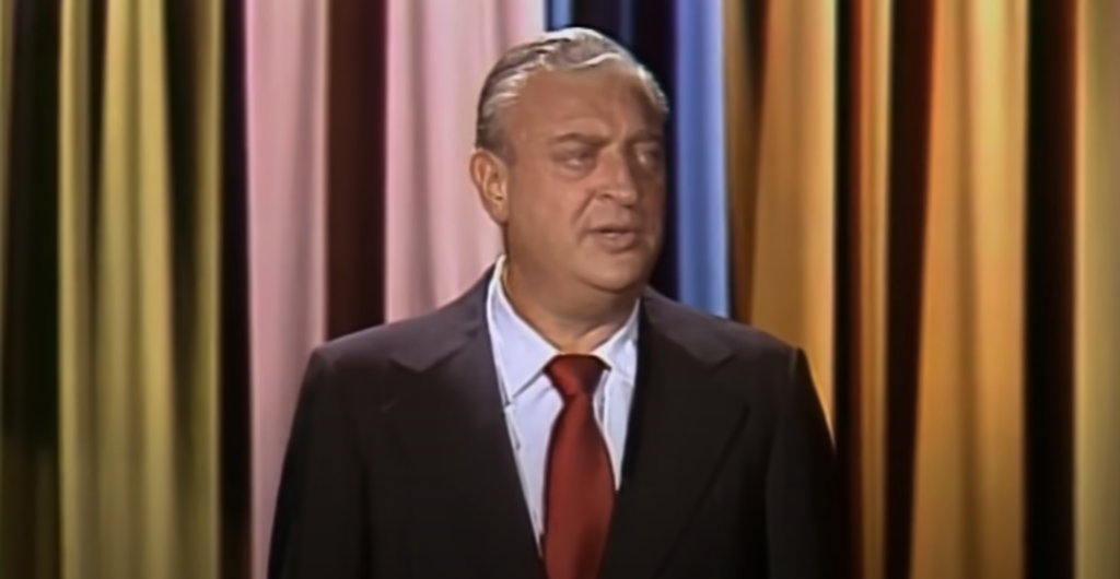 A man in a dark suit and red tie stands in front of a colorful curtain backdrop, looking to the side with a neutral expression. The background features vertical stripes in various colors, including pink, blue, and yellow.