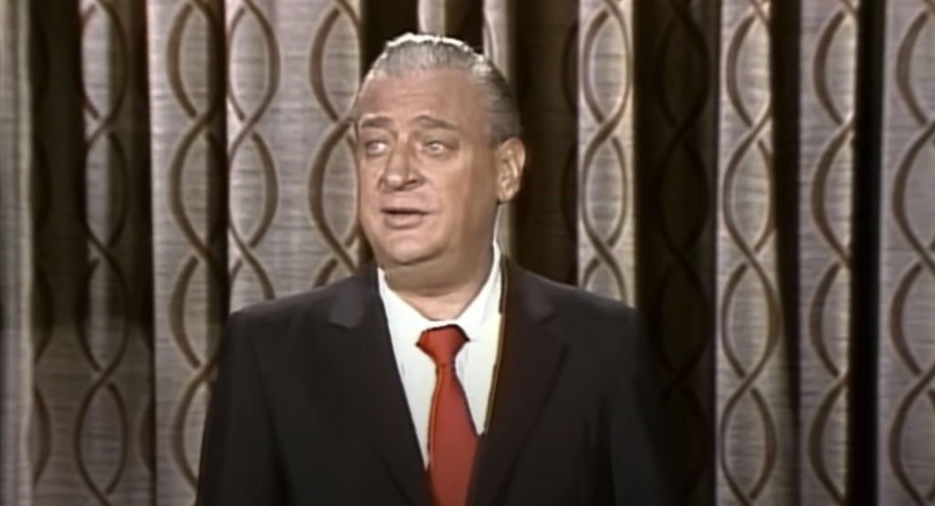 A man with slicked-back grey hair, wearing a dark suit, white shirt, and red tie, stands in front of a patterned curtain, appearing to speak or perform.