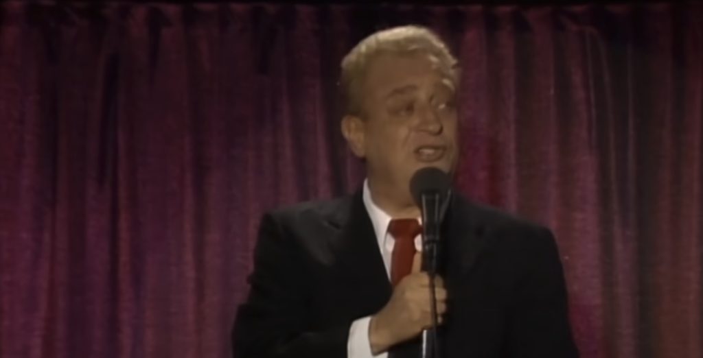 A person wearing a black suit, white shirt, and red tie is standing on a stage with a dark curtain backdrop, holding a microphone and appearing to speak or perform.