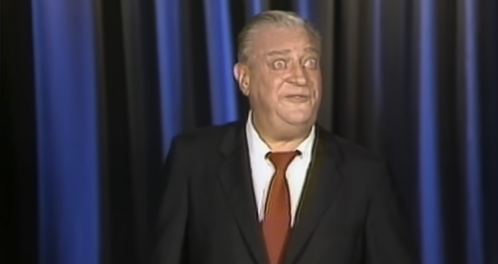 A man with gray hair, large eyes, and an expressive face is wearing a black suit, white shirt, and red tie. He is standing in front of a black and blue curtain and appears to be speaking or performing.