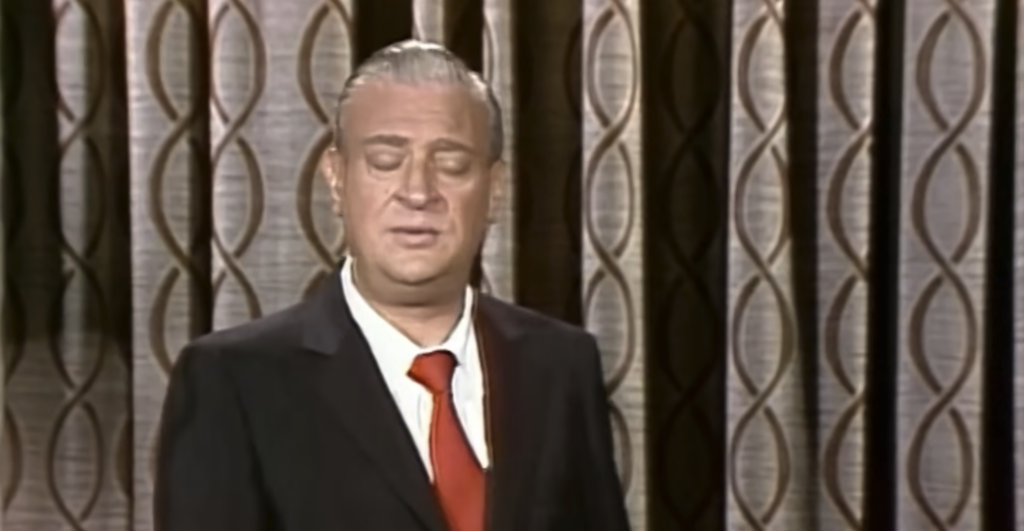 A man in a suit with a red tie stands in front of a curtain with a geometric pattern. His eyes are closed, and he appears to be mid-speech or performing on stage. The curtain features an overlapping oval design.