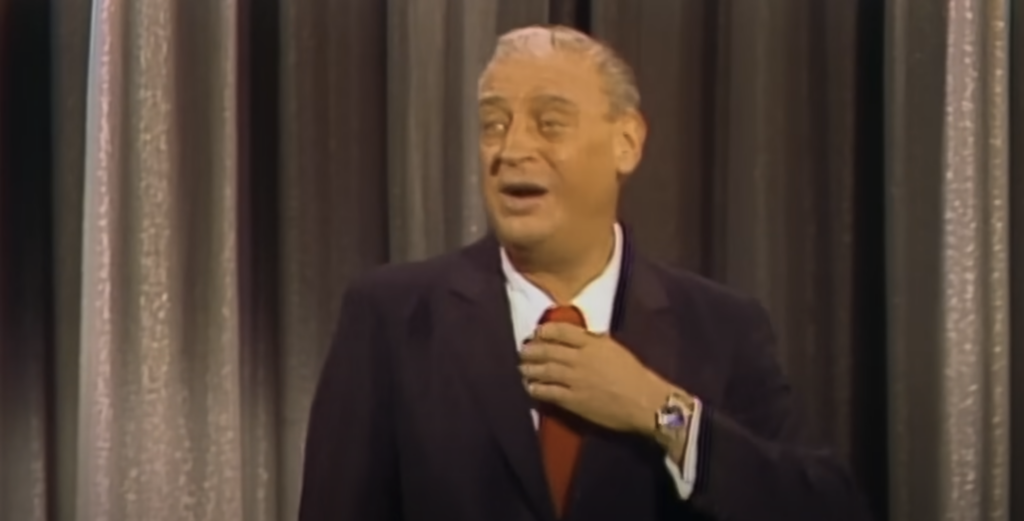 A person with grey hair is standing in front of a curtain backdrop, wearing a dark suit, white shirt, and red tie. The person appears to be mid-speech, gesturing with one hand placed near their neck, and wearing a wristwatch on the other hand.