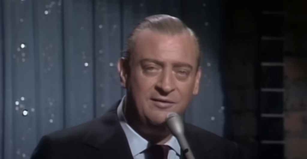 A man with neatly combed back hair and a serious expression is speaking into a microphone. He is dressed in a dark suit and tie. The background features a curtain with small, sparkling lights.