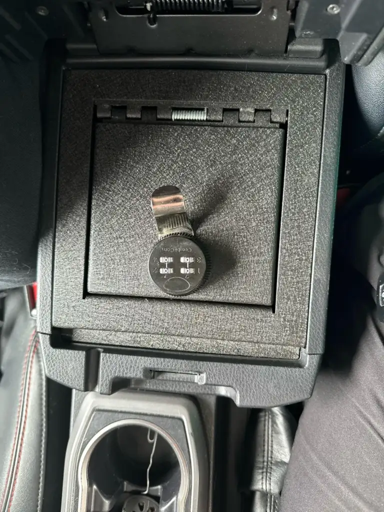 A car center console is open, revealing a lockable compartment with a combination lock and latch. The surrounding area includes cup holders and a black interior with red stitching. The compartment has a textured black surface.