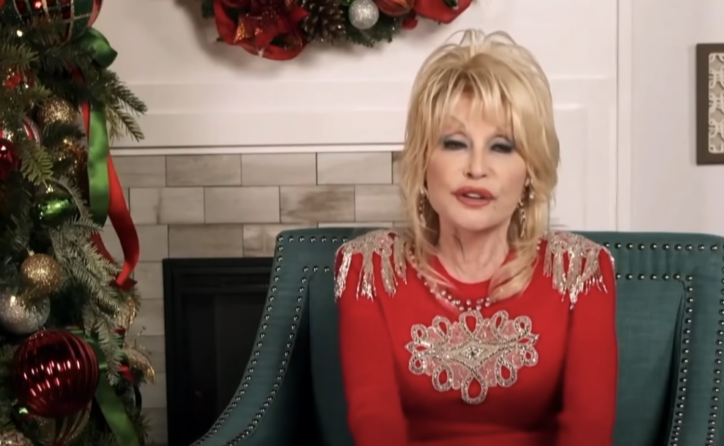 Dolly Parton talking while sitting next to a Christmas tree. 