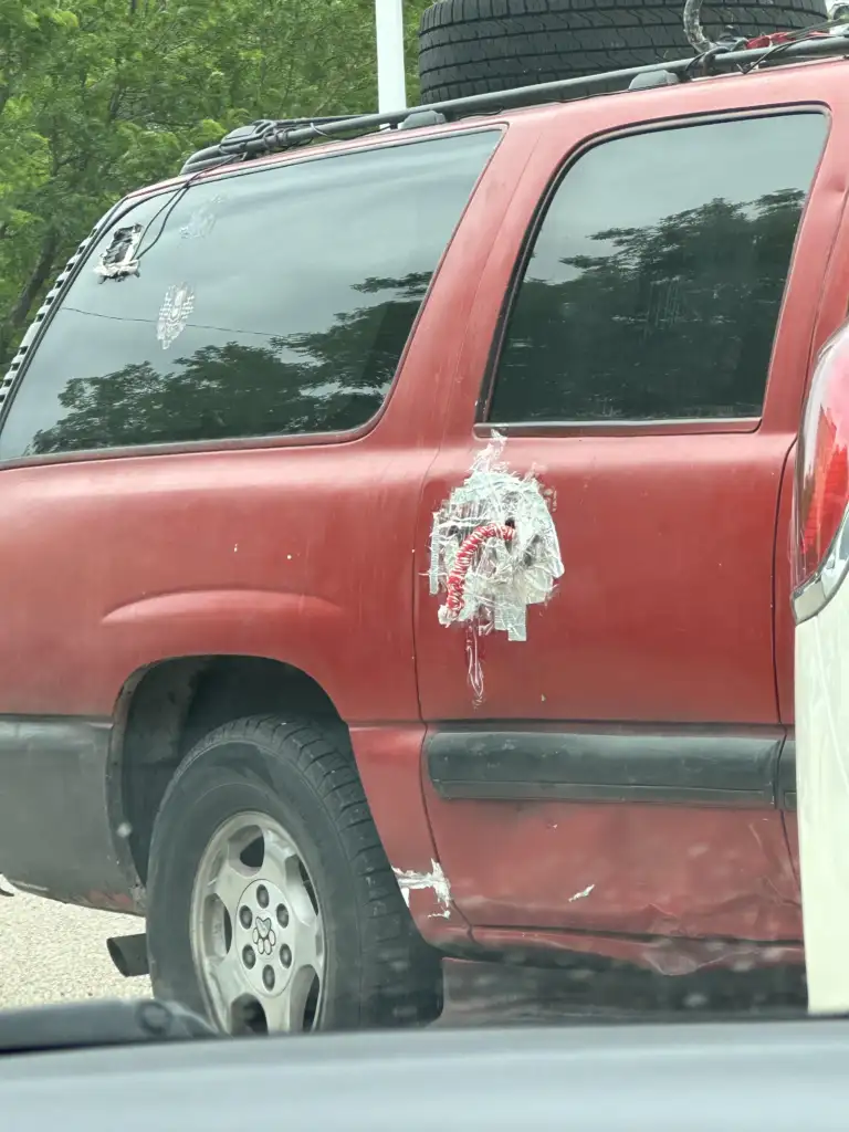 A red SUV with a damaged rear door on the driver's side, temporarily patched with clear packing tape and red fabric. The vehicle has visible scratches and dents, and a mounted spare tire on the roof. Surrounding trees are reflected in the vehicle's windows.