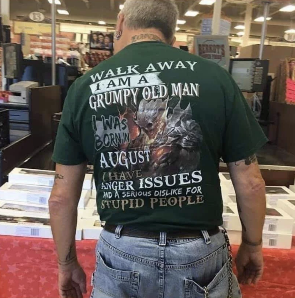 A man with short gray hair, tattoos on his arms, and a chain hanging from his belt is wearing a dark green T-shirt. The back of the shirt reads, "Walk away I am a grumpy old man I was born in August I have anger issues and a serious dislike for stupid people.