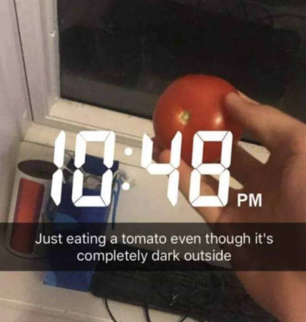 A hand holding a tomato is seen in front of a window showing darkness outside. The image has a digital timestamp reading 10:48 PM and a caption that reads, "Just eating a tomato even though it's completely dark outside.