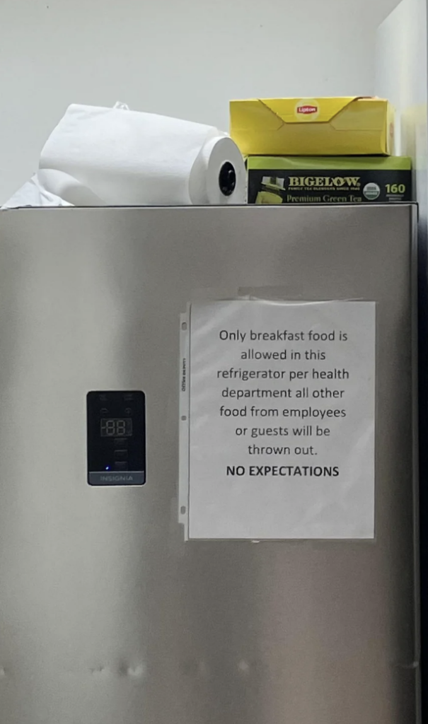 A refrigerator with a sign taped to the door reads, "Only breakfast food is allowed in this refrigerator per health department all other food from employees or guests will be thrown out. NO EXPECTATIONS." On top of the fridge are paper towels and tea boxes.