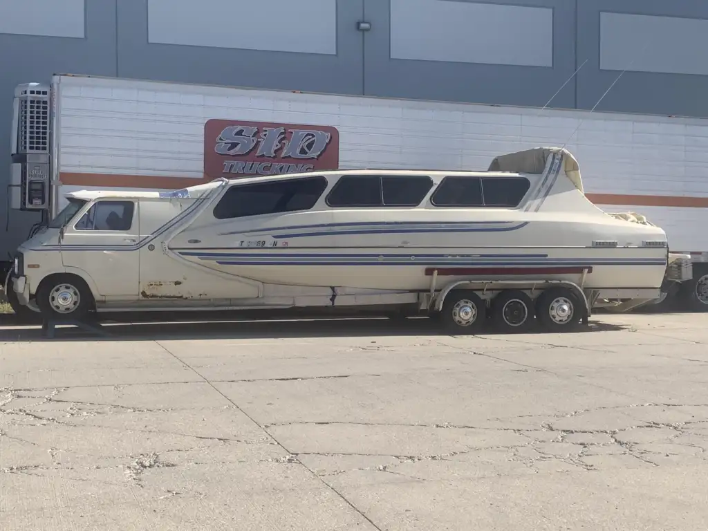 A heavily customized vehicle sits parked on the street, combining elements of a van and a boat. It has a boat-like upper structure seamlessly integrated with the body of a van and is mounted on a triple-axle automobile platform. A truck and warehouse are in the background.