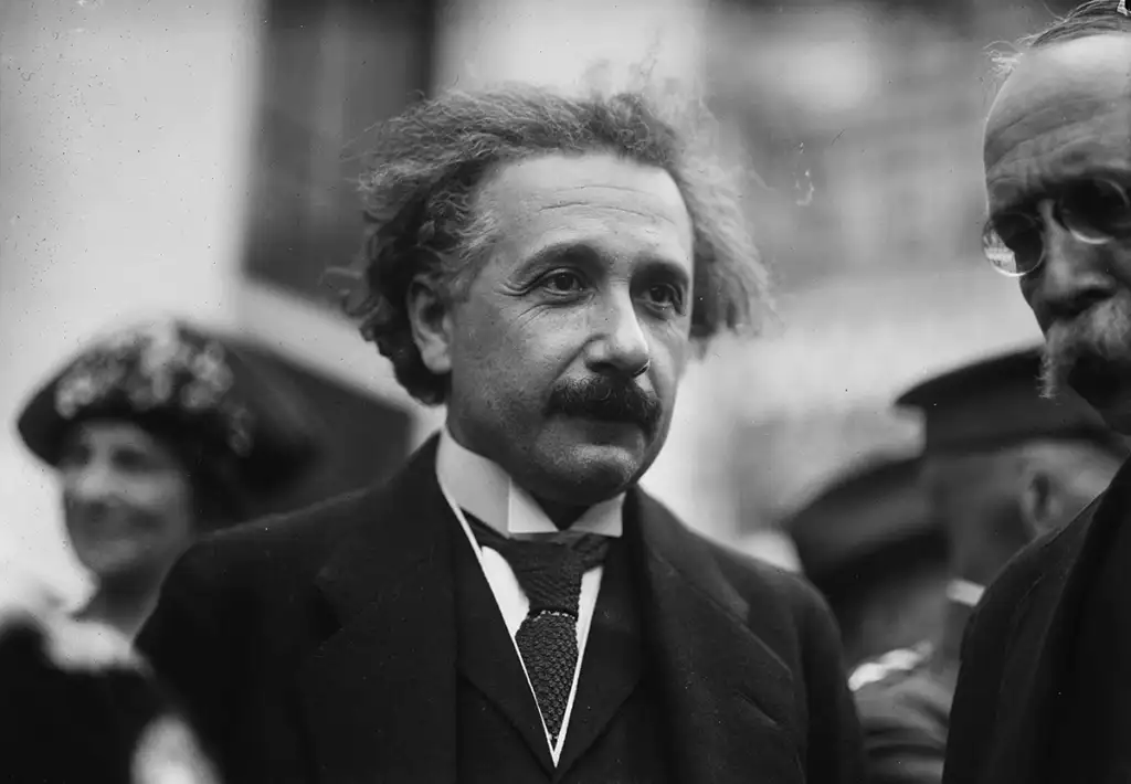 A black-and-white photograph of a middle-aged man with wild, curly hair and a mustache, wearing a suit and tie. He appears to be engaged in conversation with another man partially visible to the right. A blurred figure in the background suggests a crowd.