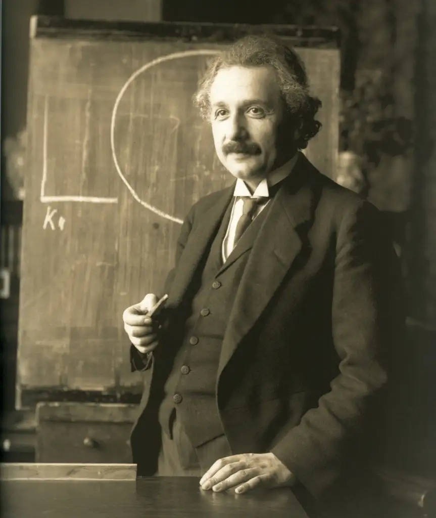 A black-and-white photograph of a man with a mustache and wild hair, wearing a dark three-piece suit and tie. He stands in front of a chalkboard with a diagram drawn on it, holding a piece of chalk in his right hand while looking at the camera.