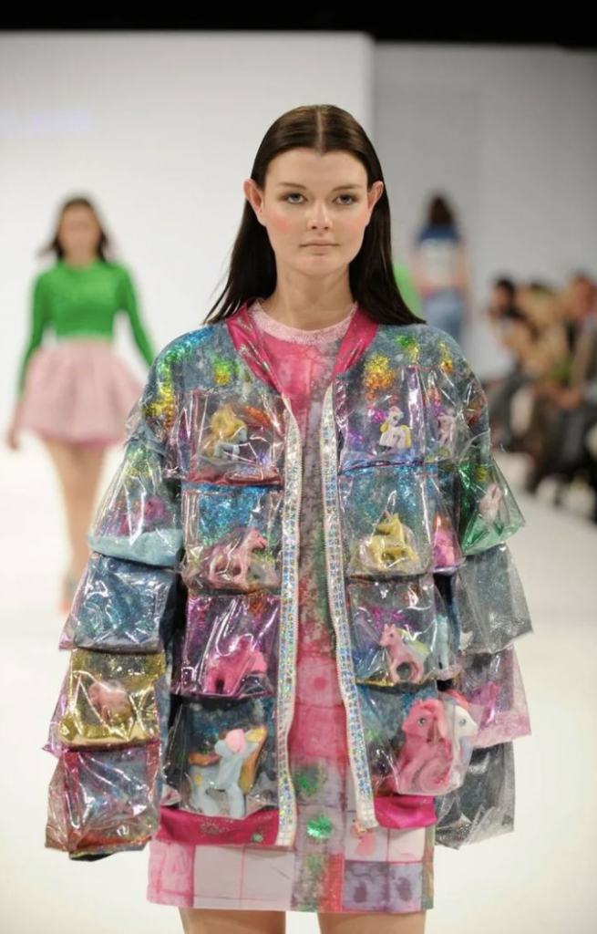 A model on a runway wearing a transparent, multicolored, glittery jacket with various small plush toys in its pockets. The outfit also includes a pink patterned dress underneath. Another model in a green top and pink skirt is visible in the background.