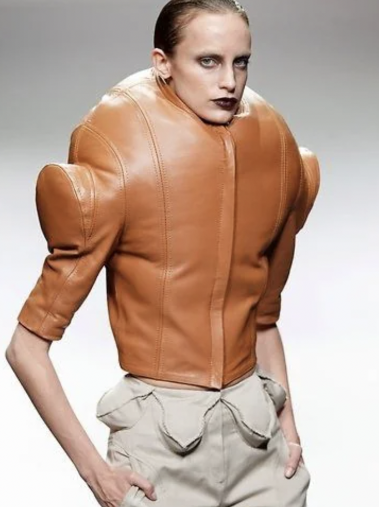 A model with slicked-back hair wears a unique, structured brown leather jacket with exaggerated shoulders and a high collar, paired with beige pants that have ruffled details at the waist. The background is a simple grey gradient.