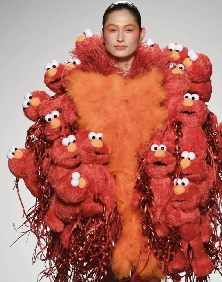 A person is wearing a vibrant, avant-garde outfit covered in numerous plush Elmo toys and red tinsel. The upper part of the outfit features an orange, fluffy heart-shaped design, while the individual's face displays bright, colorful makeup.