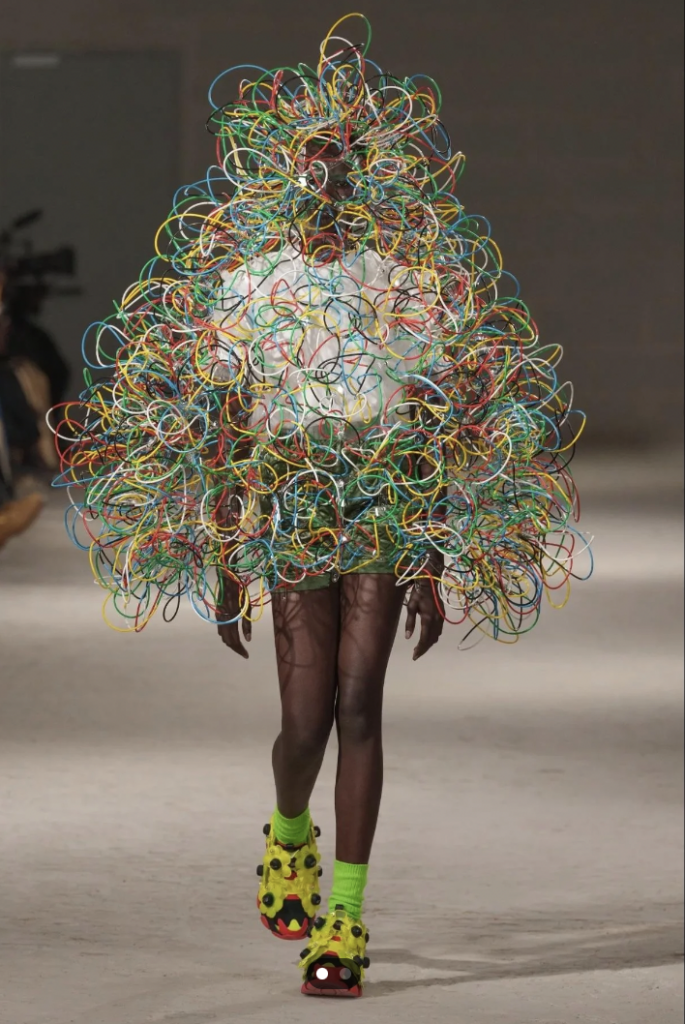 A model walks the runway wearing an avant-garde outfit made of colorful, tangled wires enveloping their entire body, creating a spherical shape around them. They are also wearing bright green socks and yellow, fuzzy shoes with black accents.