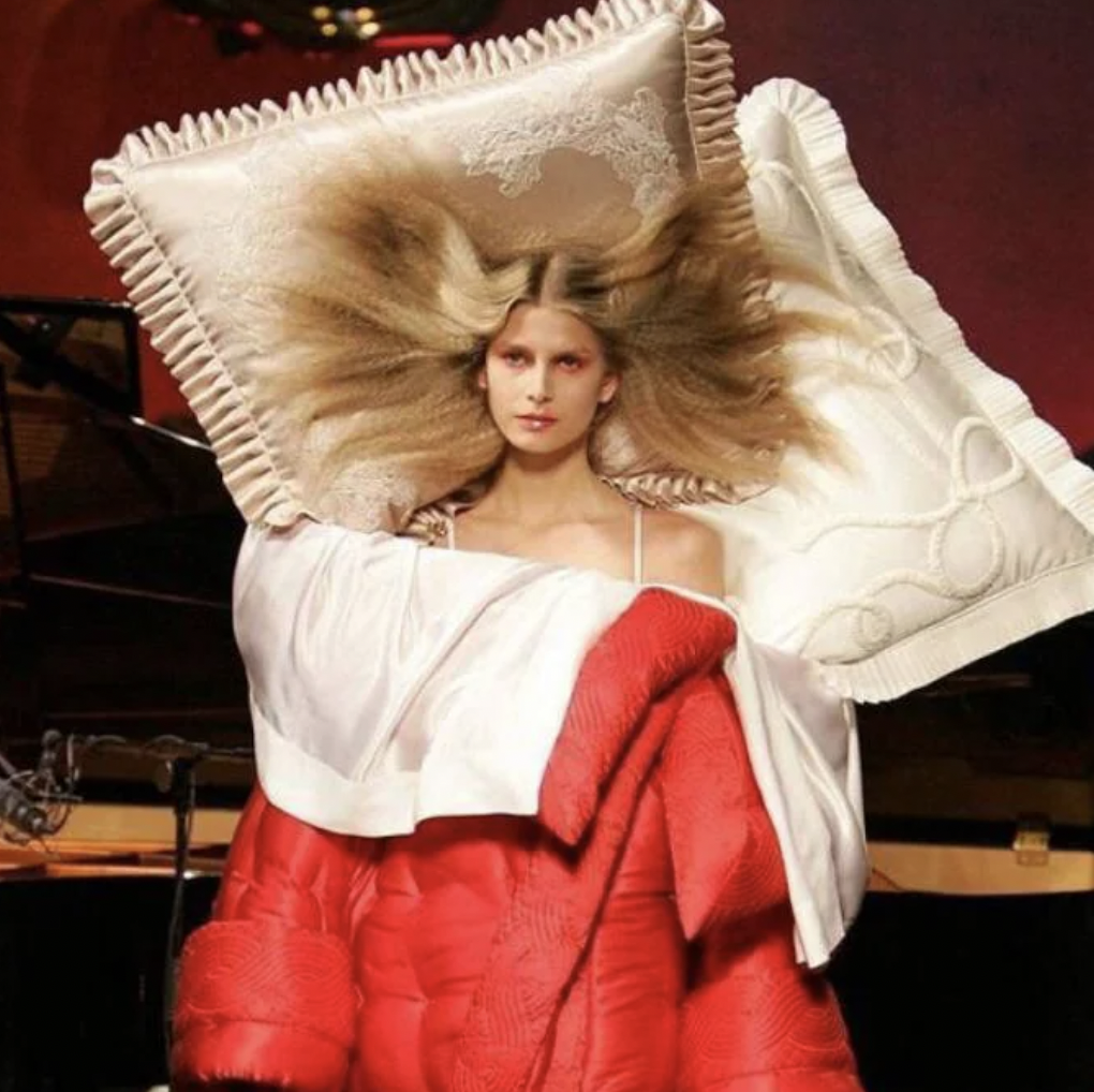 A woman on a runway wearing an avant-garde fashion outfit that resembles a bed. Her outfit includes two pillows around her head and a large red blanket draped over her shoulders. Her hair is styled to blend into the pillows, creating a unique and surreal look.