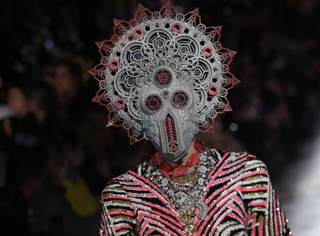 A model is wearing an elaborate headpiece resembling a tribal mask with intricate geometric patterns in silver and red colors. The outfit includes a black, white, and pink striped top adorned with multiple layers of necklaces.