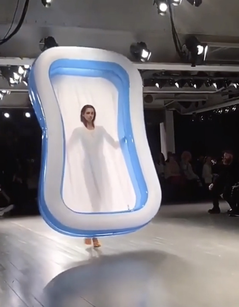 A model walks down a runway in a fashion show, wearing a unique outfit shaped like a large inflatable pool. The outfit frames the model and is blue and white. The event is lit with overhead stage lighting and a seated audience is visible in the background.