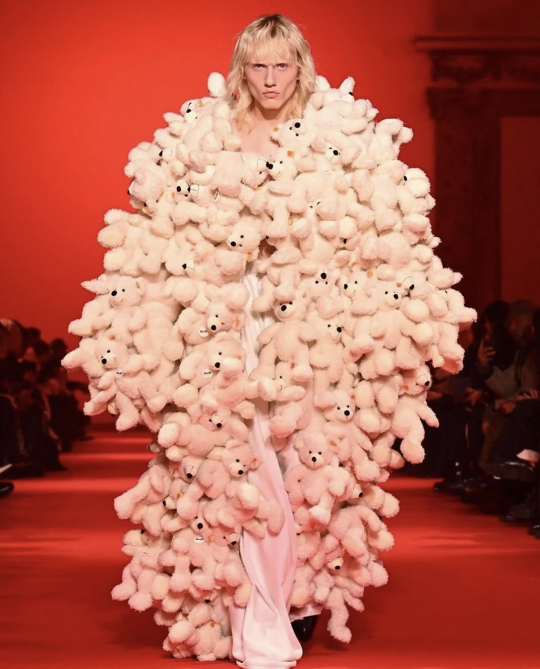 A model on a fashion runway wears an avant-garde outfit covered entirely in small, pale teddy bears. The background is lit with a red hue, and an audience is visible on the sides. The model has long blonde hair and an intense expression.