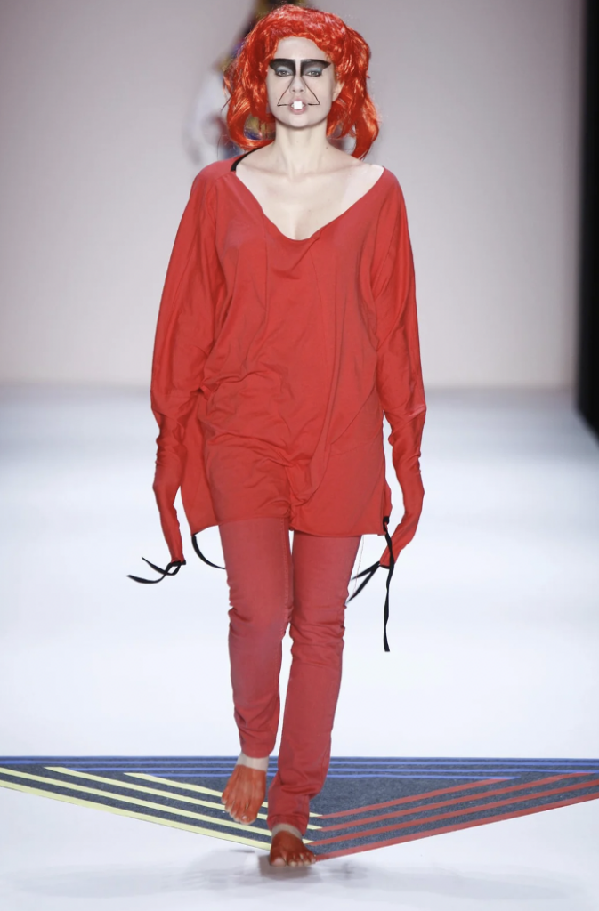 A person walks down a runway wearing a bold red outfit consisting of a loose shirt, pants, and gloves, paired with a vibrant orange wig. The individual has distinctive face makeup featuring a black geometric design around the eyes and mouth.