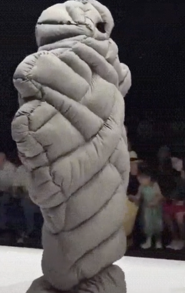A person walks down a runway dressed in an oversized, puffy, grey costume that resembles a caterpillar or segmented worm. The audience on both sides watches as the unique outfit is showcased.