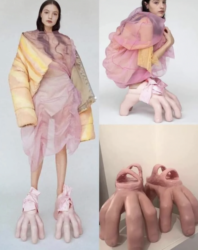 A fashion model wears a sheer pink outfit paired with an oversized yellow sleeve. The model also dons unusual shoes resembling large, fleshy human fingers. Close-up shots of the shoes highlight their realistic, hand-like appearance.