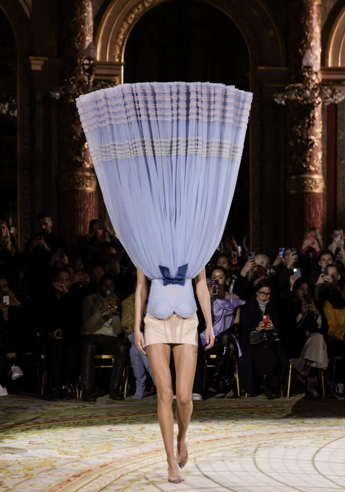 A fashion model walks down a runway wearing a unique outfit consisting of a light blue curtain-like headdress that covers the upper body, paired with beige high-waisted shorts. Spectators in dark clothing watch and take photos in an ornate, gilded room.