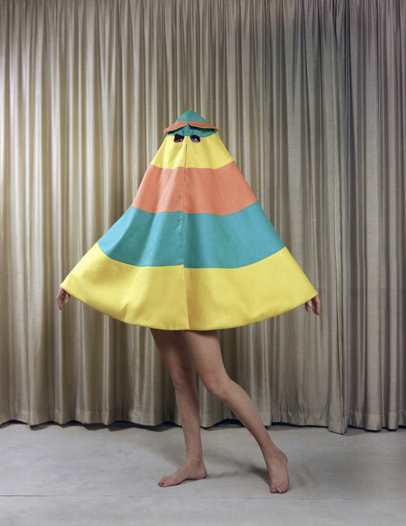 A person stands in a room wearing a colorful, cone-shaped outfit with horizontal stripes in yellow, orange, teal, and blue. Their bare legs and feet are visible below the outfit, and they have small eye holes near the top. Beige curtains hang in the background.