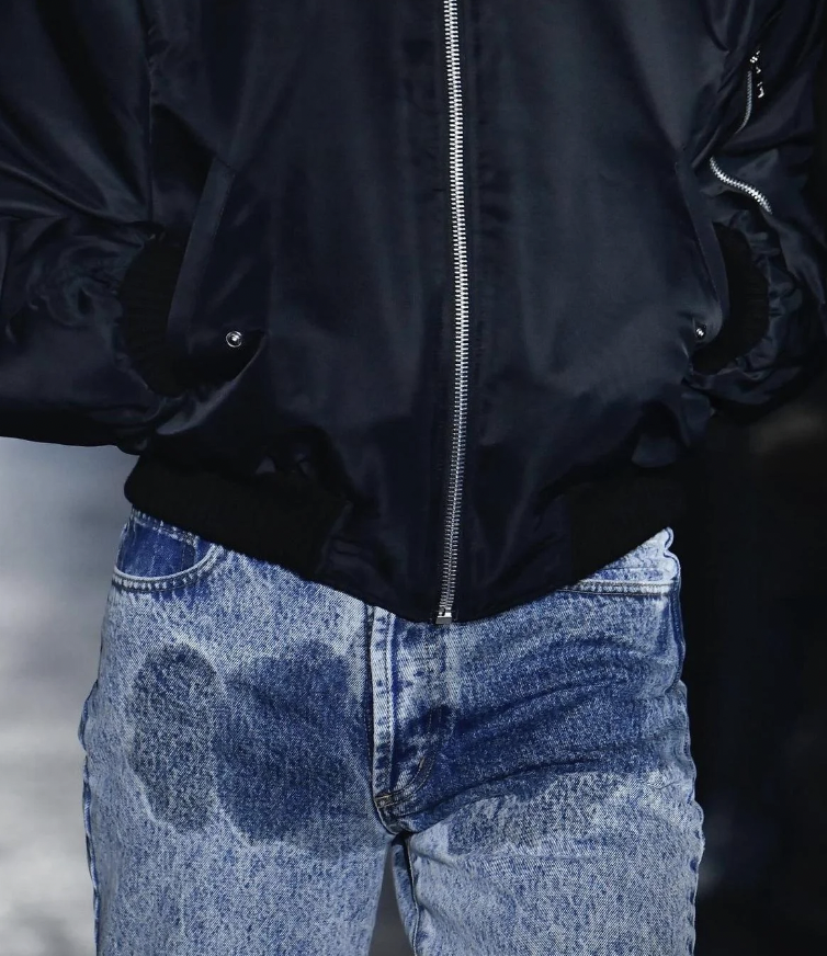 A person wearing a black jacket with a silver zipper and dark blue jeans. The jeans have a distinct pattern that resembles water stains across the front, creating a unique, weathered appearance. The person's hands are in the jacket pockets.