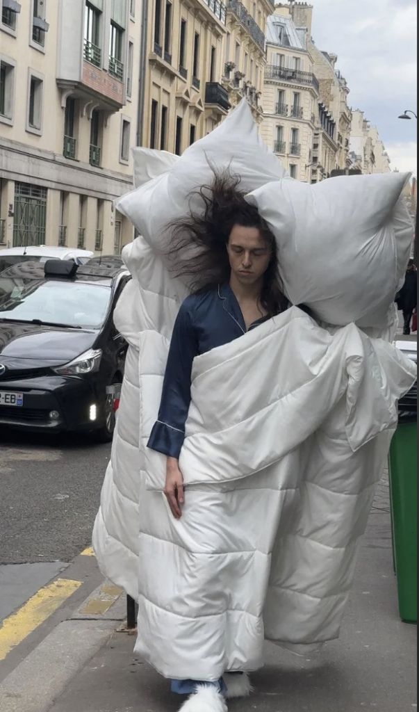 A person with long dark hair, wearing dark blue pajamas, walks on a city sidewalk wrapped in a white comforter. Large white pillows are balanced on their shoulders. The street is lined with buildings and parked cars.