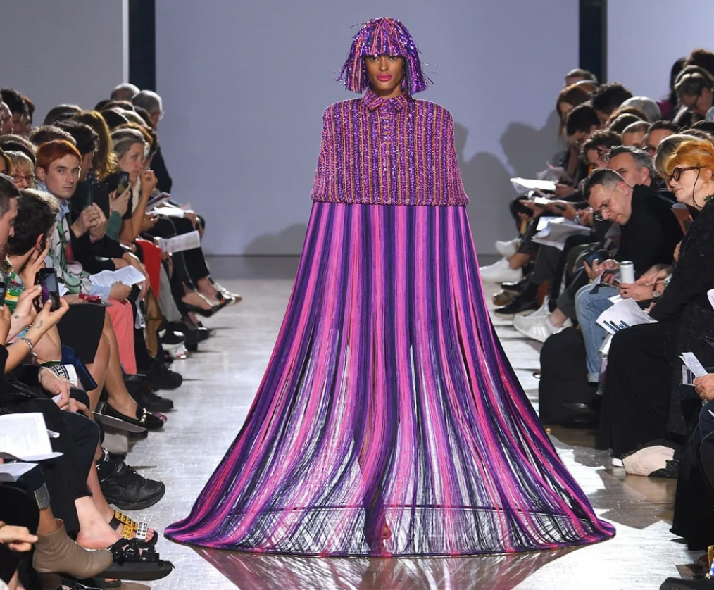 A person walks down a runway wearing a vibrant, floor-length, purple and pink striped outfit with a matching headpiece. The audience is seated on both sides, attentively watching the fashion show. The room is well-lit, highlighting the bold attire.