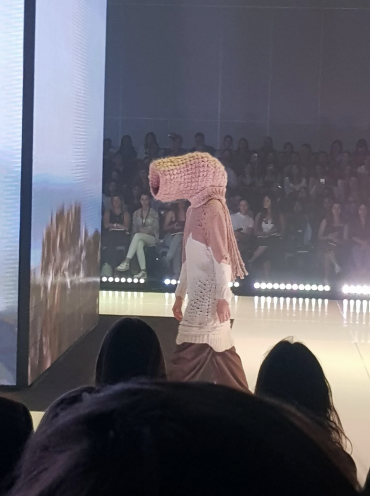 A model walks down a runway wearing a white and brown dress with a large, uniquely shaped crochet pink hood obscuring most of their head. The audience is seated on both sides of the catwalk, with bright stage lights illuminating the scene.