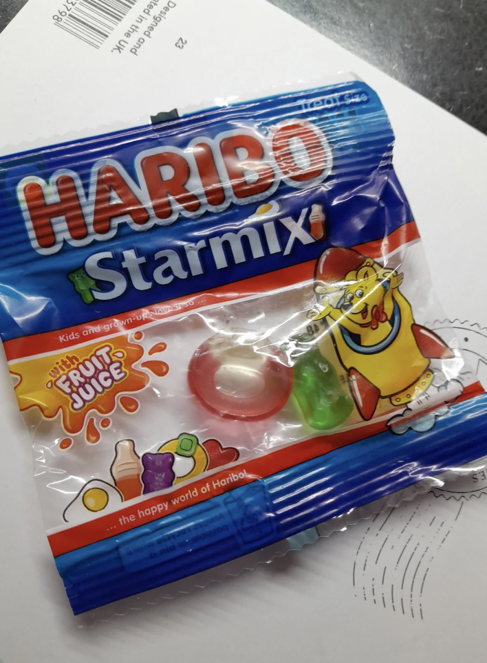 A partially empty packet of Haribo Starmix candies lies on a white surface. The packaging is blue, with the Haribo logo at the top and an illustration of various gummy candies. Visible through the packaging are a red-and-white gummy ring, a green gummy candy, and an empty space where other candies have been removed.