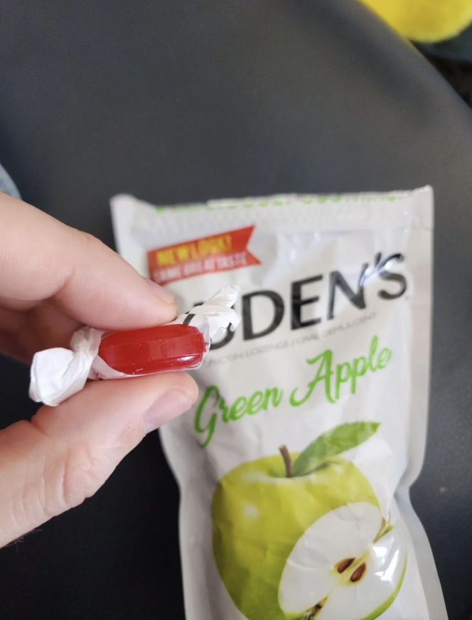 A hand holds a wrapped red lozenge in front of a partially opened white package labeled "Fisherman's Friend Green Apple." The package shows an image of a green apple with a bite taken out of it. The text "new flavor" is visible at the top.