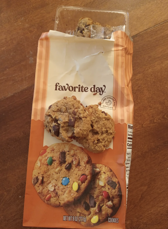 Image of a partially opened package of Favorite Day cookies on a wooden surface. The package shows cookies with chocolate chunks and colorful candy-coated pieces. Some cookies are visible inside the opened top of the package.