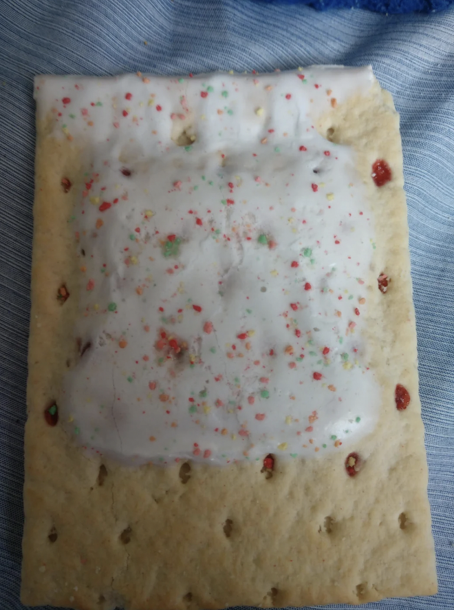 A frosted rectangular pastry with white icing and colorful sprinkles, resembling a Pop-Tart, is placed on a light blue fabric. The exposed edges show glimpses of red filling inside.