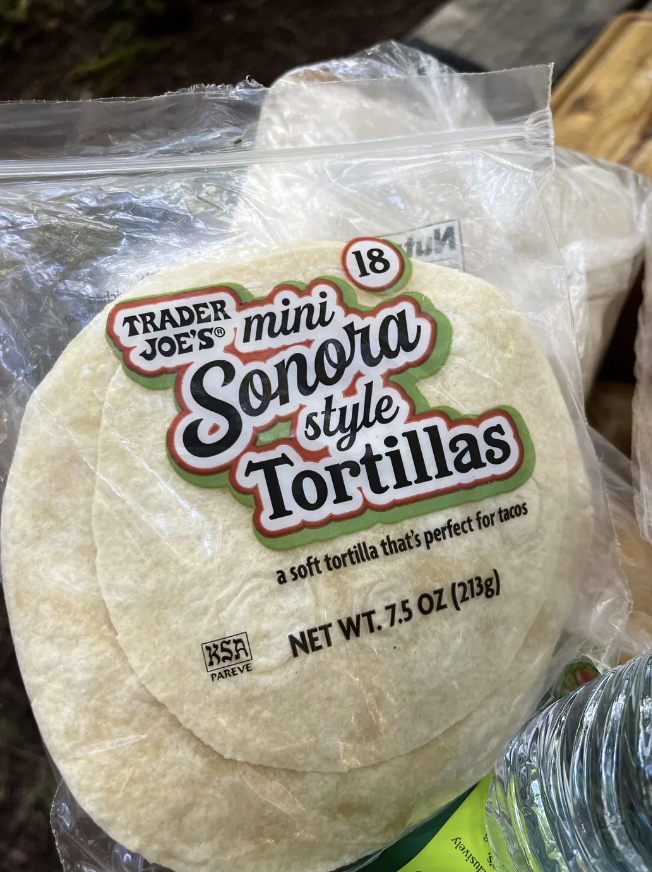 A package of Trader Joe's Mini Sonora Style Tortillas is shown. The clear plastic bag displays 18 tortillas inside, described as "a soft tortilla that's perfect for tacos." The net weight is 7.5 ounces (213 grams), and a "KSA Pareve" certification is visible.