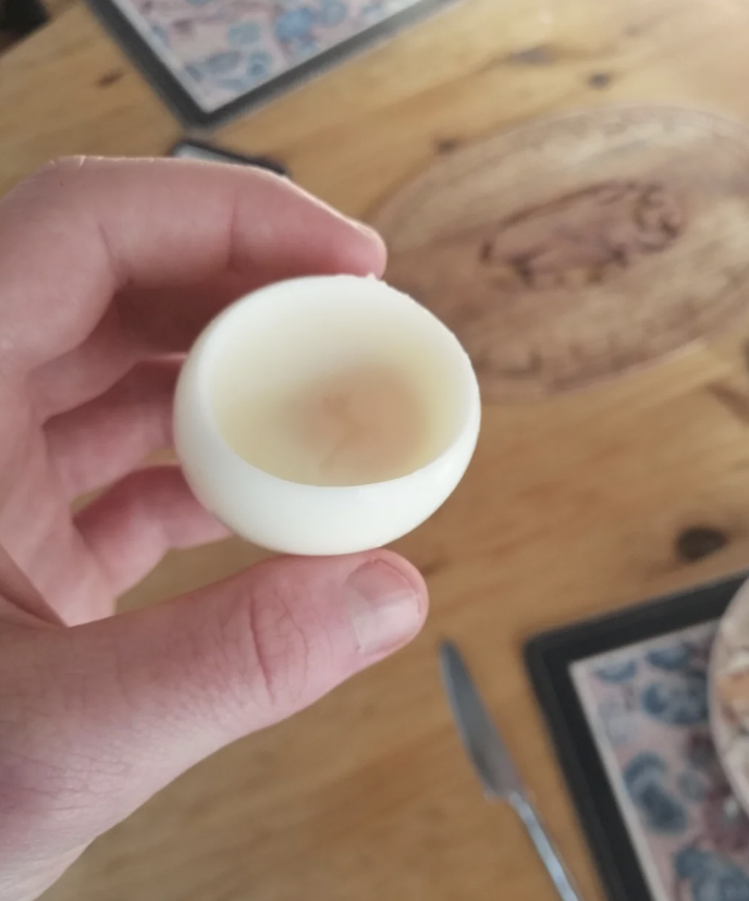 A hand holding a peeled hard-boiled egg with a small indentation where the yolk has been removed. The background shows a wooden table with placemats and a knife.