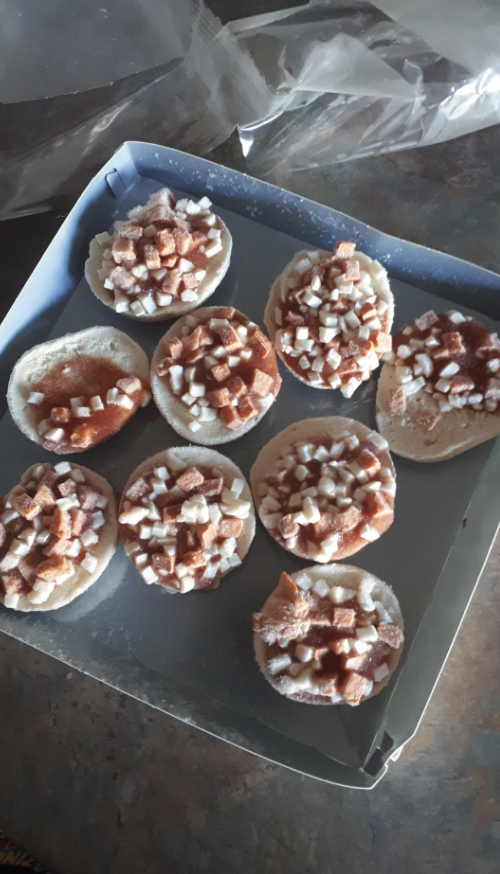 A metal tray holds nine unbaked pastries topped with small diced apples and a sprinkle of cinnamon sugar, likely prepared for baking. The pastries are arranged in rows on a kitchen countertop.