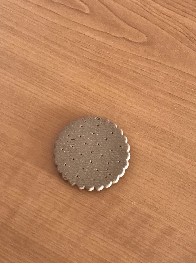 A single round chocolate biscuit with a pattern of small holes is placed on a light brown wooden surface. The edges of the biscuit are scalloped.