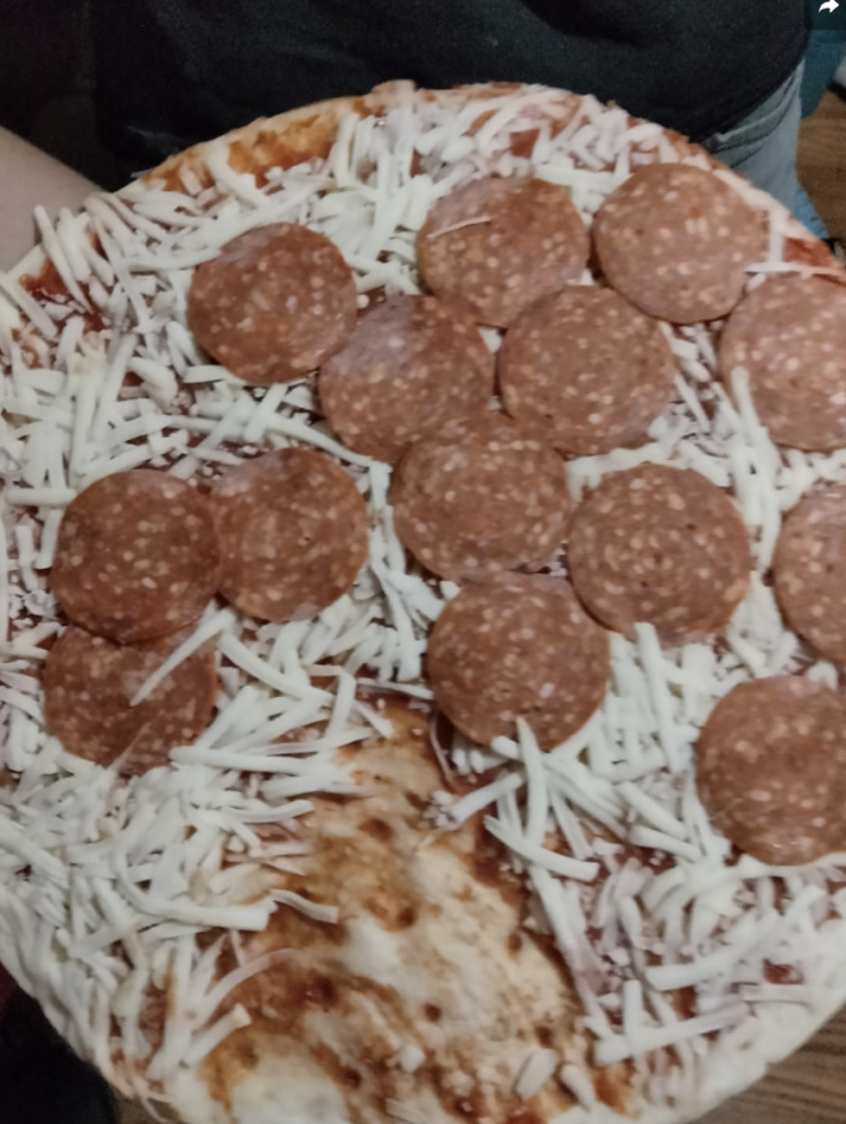 A close-up of an uncooked pizza with a layer of shredded mozzarella cheese and pepperoni slices on a thin crust. Some areas along the edge appear to have missing cheese. The background is slightly blurred, showing parts of a person's upper body.