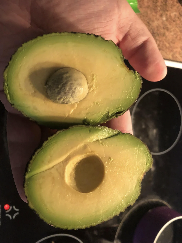 A hand holding a halved avocado with the pit still in one half and removed from the other. The ripe avocado has a dark green, slightly textured skin and smooth, bright green flesh.