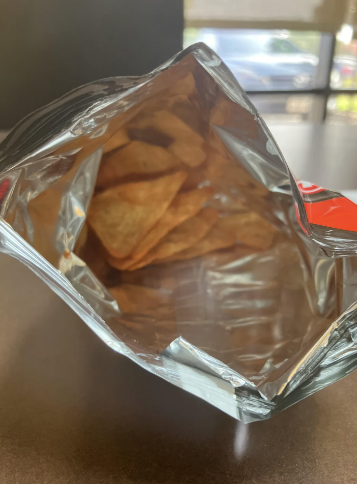 An open bag of red seasoned tortilla chips is shown, viewed from above. The bag is partially filled with the chips, displaying their triangular shape and vibrant seasoning. The background is slightly blurred, suggesting an indoor setting.