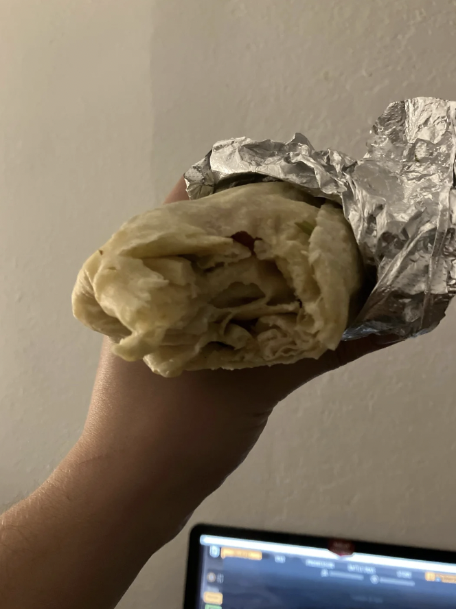 A close-up of a hand holding a partially wrapped burrito in foil. The burrito appears to be filled with ingredients like meat and vegetables. The background is a blank wall, and part of a computer screen is visible at the bottom of the image.