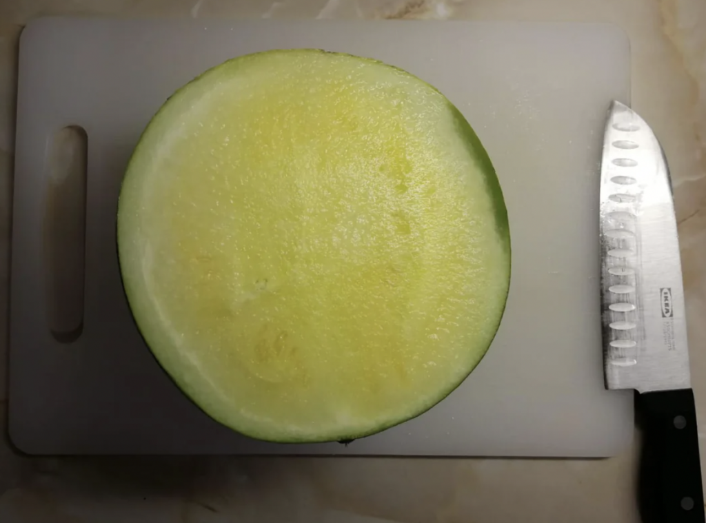 A half-cut yellow watermelon is placed on a white cutting board. A silver knife with a black handle lies to the right of the watermelon. The surface underneath the cutting board has a beige pattern.