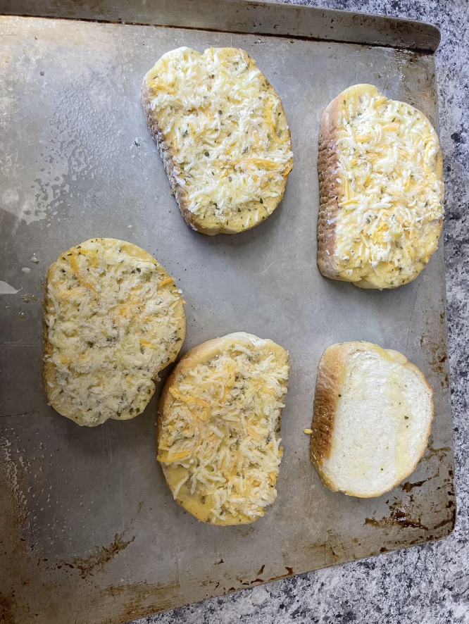 An old baking sheet with five slices of bread topped with shredded cheese sits on a gray countertop, ready for baking. Four slices have cheese evenly spread, while one slice has cheese only on the right edge. The bread appears to be seasoned with black pepper.